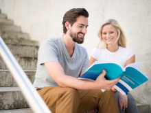Bildtyp: Foto; Beschreibung: Zwei Personen sitzen zusammen auf einer Treppe und schauen sich gemeinsam ein Handbuch an.